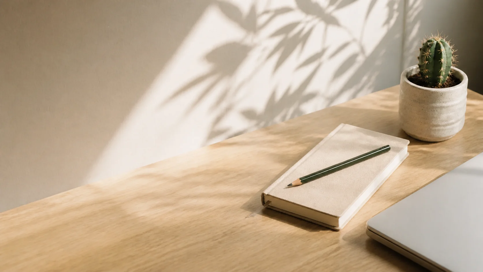 A quiet desk with a notebook, pencil, small cactus, and closed laptop in morning light